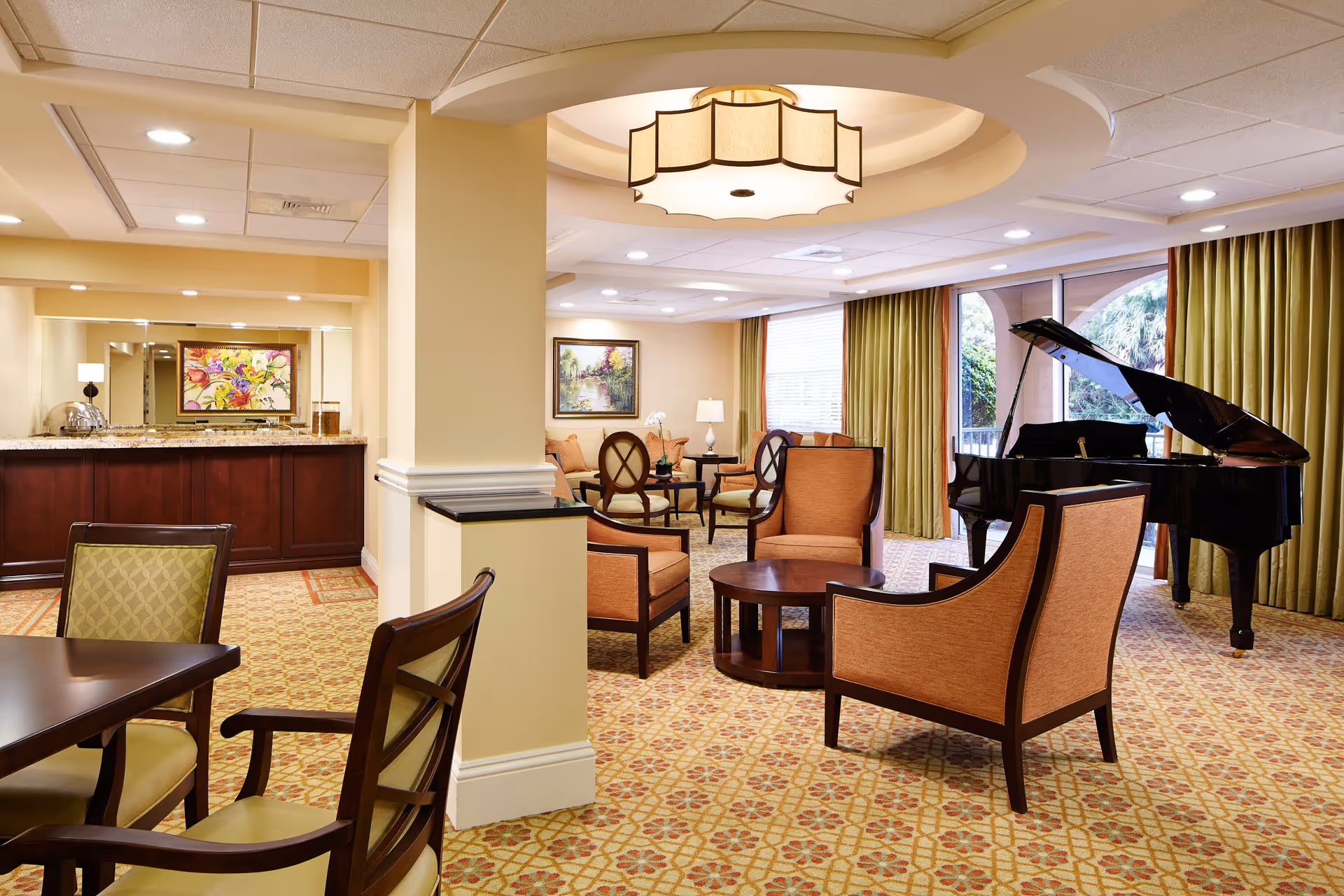 A spacious, well-lit living room area with patterned carpet, several upholstered chairs and sofas arranged around wooden coffee tables, a grand piano near large windows with green curtains, and a buffet counter with a colorful floral painting above it.