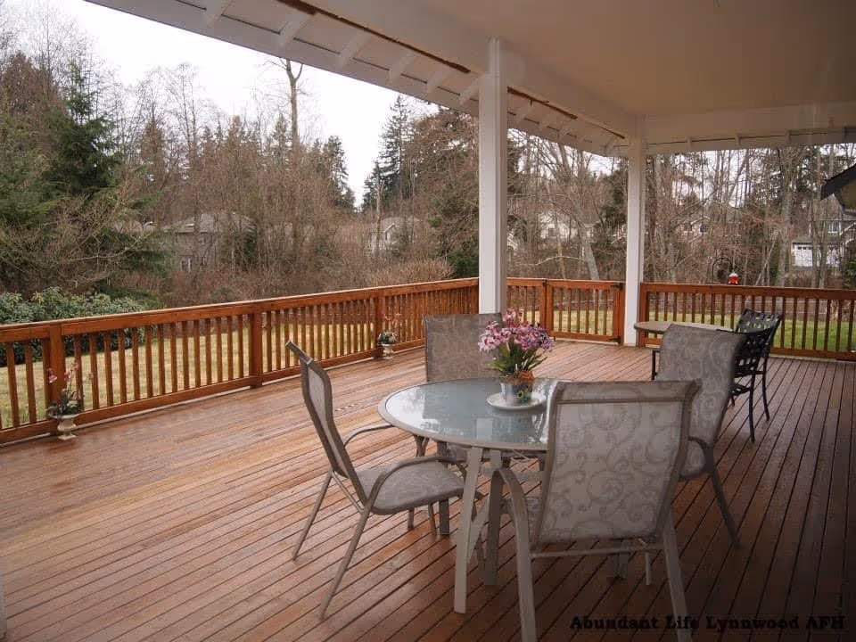 Covered wooden deck with a round glass table and four cushioned chairs. There are potted plants on the deck railing and a wooded area with trees in the background.