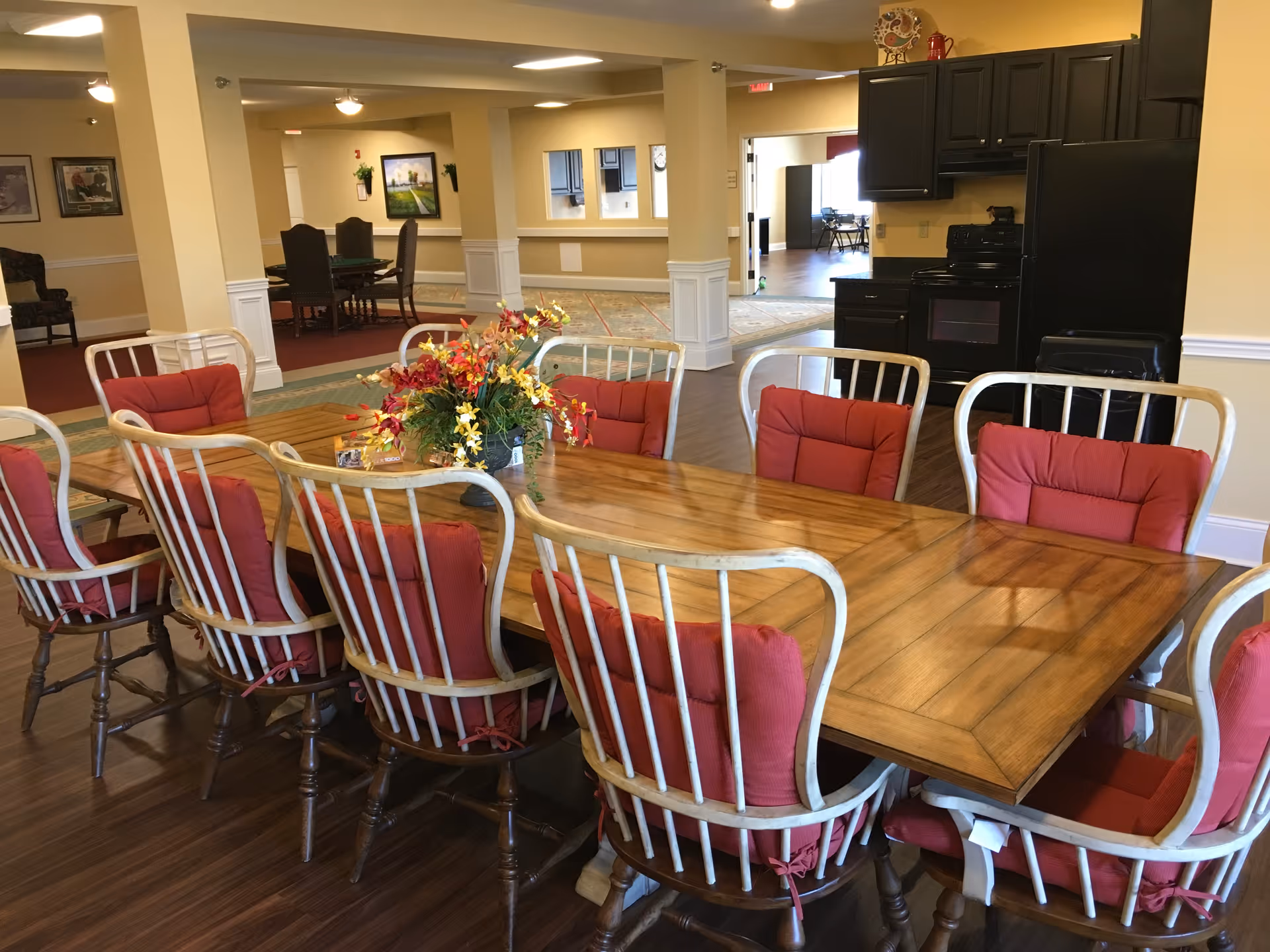 Long wooden dining table with red-cushioned chairs in a communal dining area with a kitchen visible in the background.
