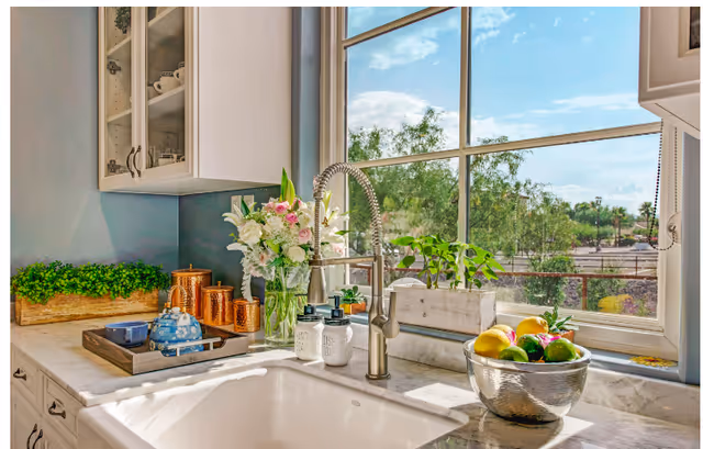 Bright kitchen sink area with a large window showing outdoor greenery. The countertop has a vase with flowers, copper containers, a tray with a teapot and cup, soap dispensers labeled 'HAND SOAP' and 'DISH SOAP', a small potted plant, and a bowl of fresh fruit including lemons and limes.