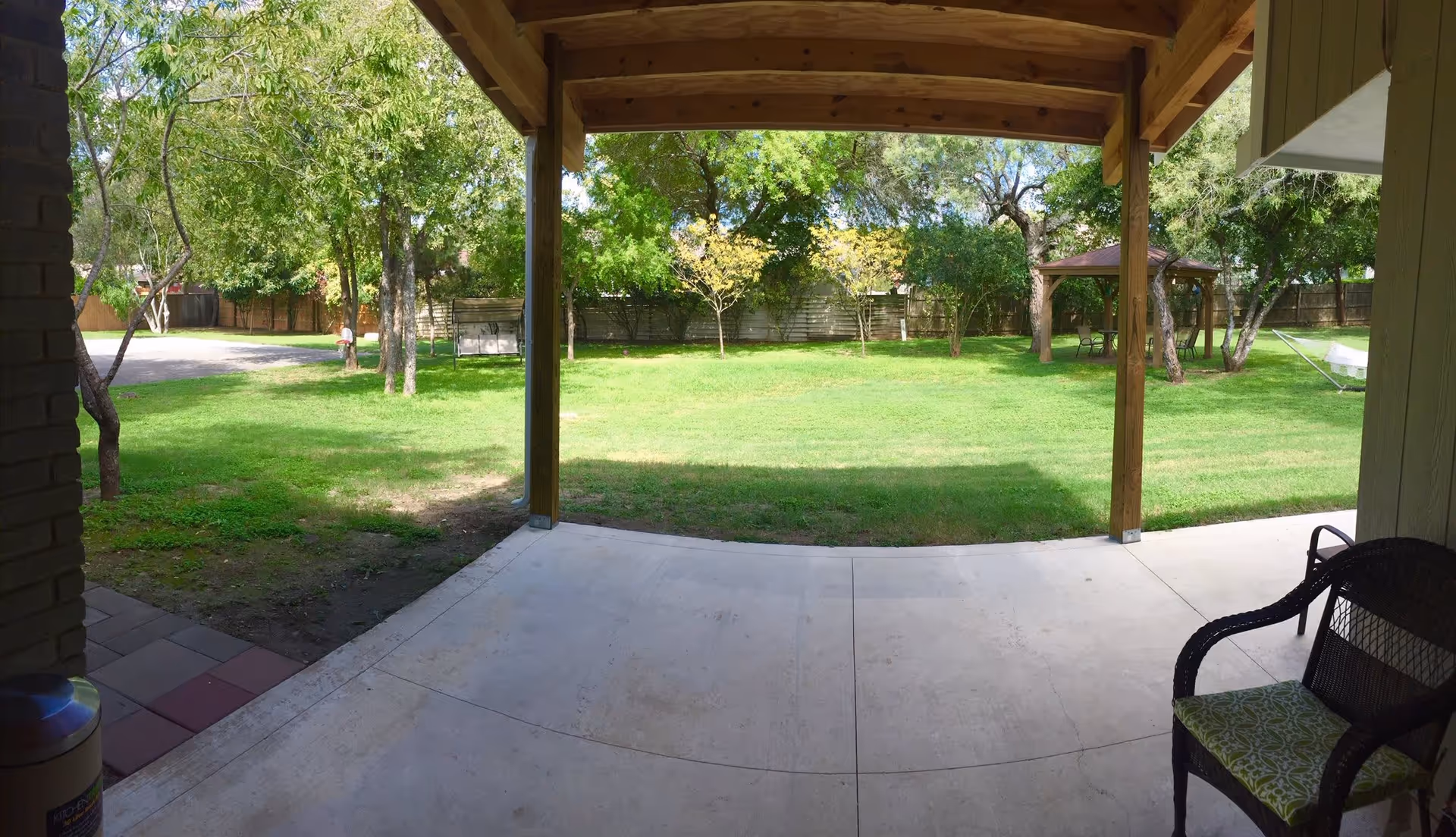 View from a covered patio looking out onto a grassy backyard with several trees, a wooden swing, a gazebo with chairs and a table, and a hammock. There is a wicker chair with a green patterned cushion on the patio.