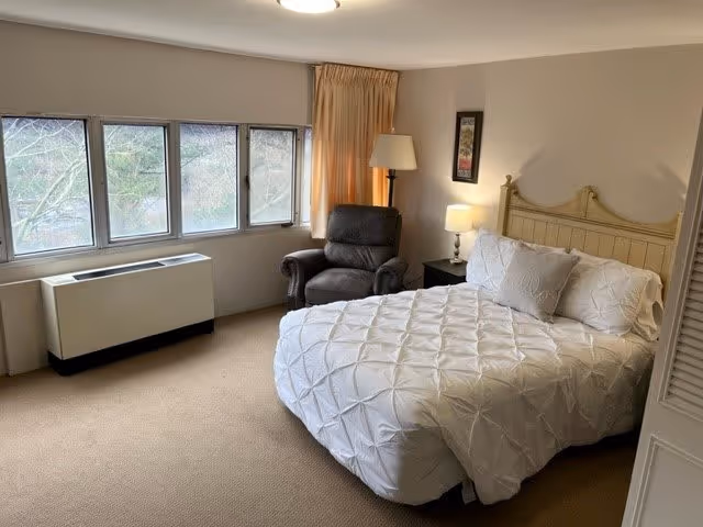 A bedroom with a large bed covered in a white textured comforter and pillows. Next to the bed is a nightstand with a lamp and a framed picture on the wall above. A dark upholstered armchair with a floor lamp behind it is positioned near a set of windows with beige curtains. The room has beige carpeting and neutral-colored walls.