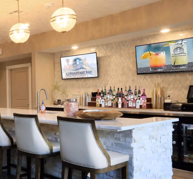 Community bar area with a marble-topped island, three bar stools, a display of bottles on the back counter and two TVs showing Chesapeake Bay graphics.