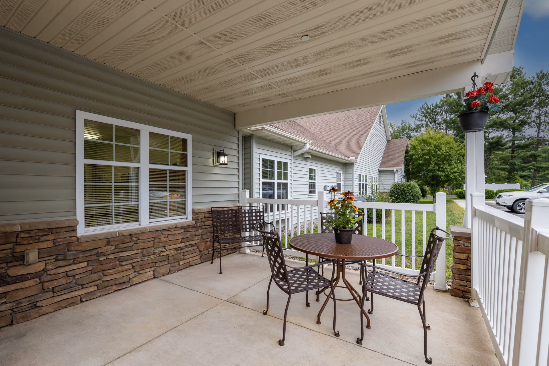 Covered outdoor patio with a round metal table, four metal chairs, potted flowers, and white railing in front of a building and lawn.