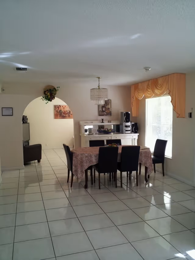 A dining room with a rectangular table covered with a floral tablecloth surrounded by six black chairs. There is a white sideboard against the wall with a stereo system on top. A window with yellow drapes allows natural light into the room. The floor is tiled with white tiles, and there is an archway leading to another room with a brown armchair visible.