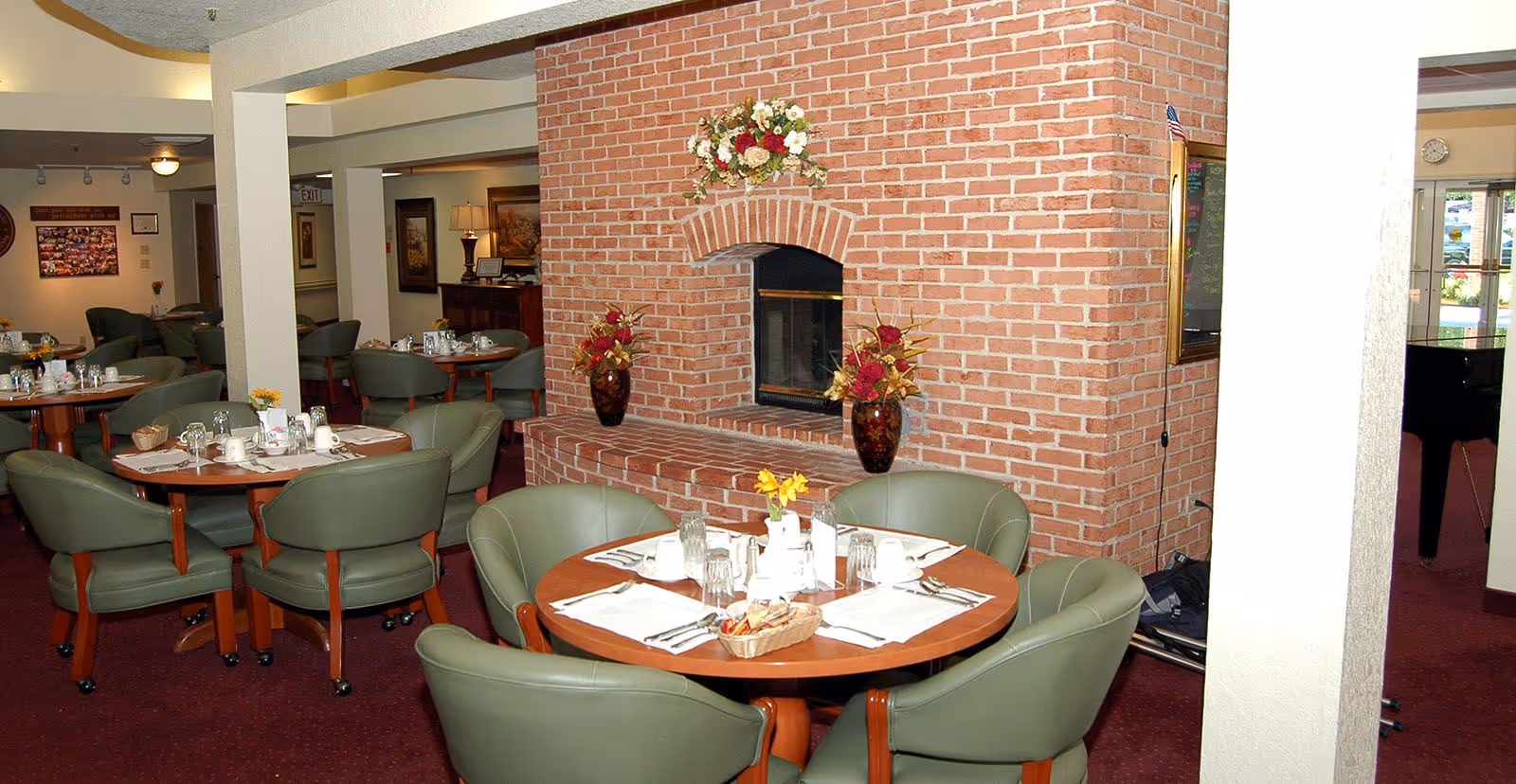 Interior view of a dining area in a senior living facility with round wooden tables set for meals, surrounded by green cushioned chairs. A brick fireplace decorated with floral arrangements is centrally located, and the room has a warm, inviting atmosphere with carpeted floors and soft lighting.