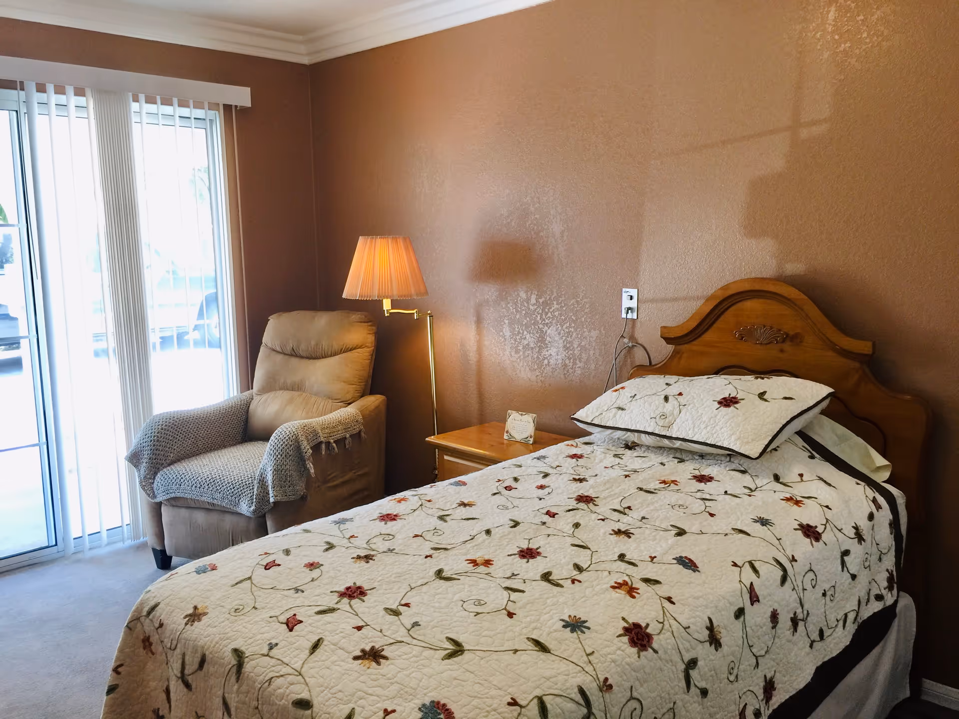 A cozy bedroom with a single bed featuring a floral quilt and matching pillow. Next to the bed is a wooden nightstand with a small decorative item and a floor lamp with a pleated shade. A comfortable armchair with a knitted throw is positioned near a sliding glass door with vertical blinds, allowing natural light to enter the room. The walls are painted a warm brown color.