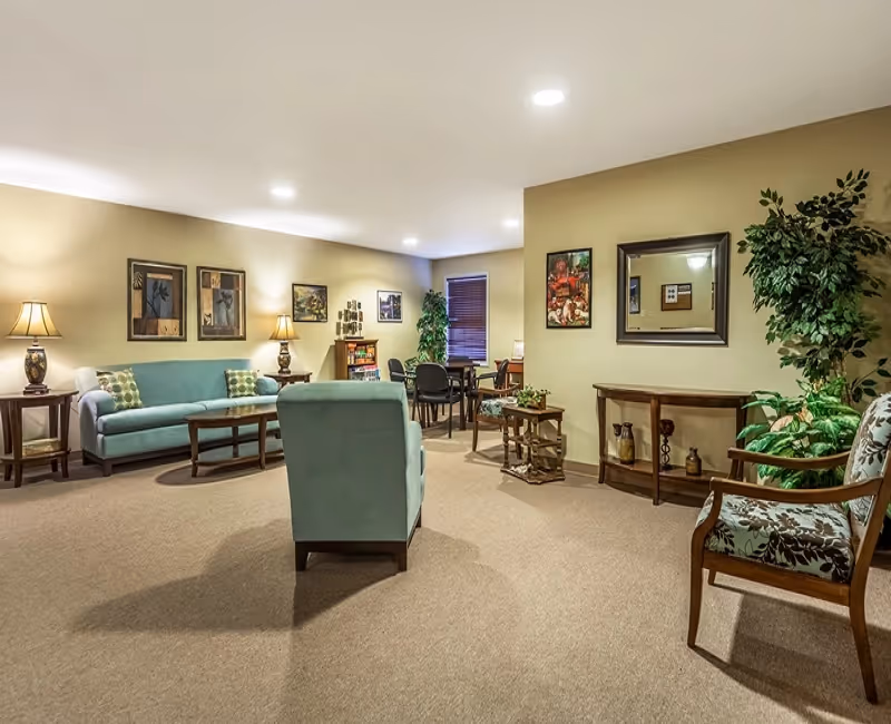 A spacious living room area in a retirement community with beige walls and carpet. The room features a light blue sofa, matching armchair, wooden coffee table, side tables with lamps, and several framed artworks on the walls. There is a small table with chairs in the background near a window with blinds, and decorative plants and a mirror on the right side.