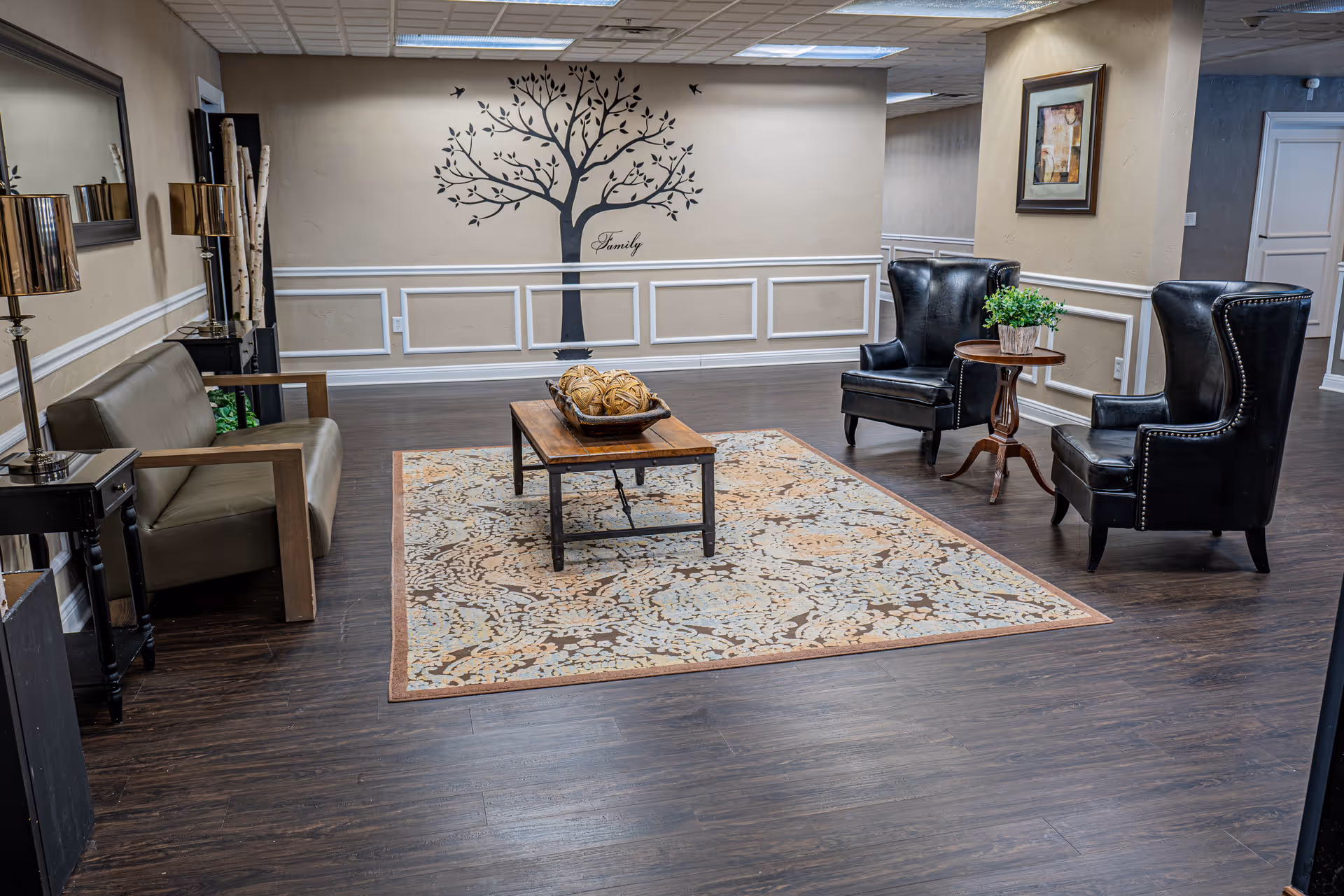 A cozy living room area with dark wood flooring, a patterned area rug, a wooden coffee table with decorative items, two black leather wingback chairs with a small round wooden table and a plant between them, and a beige sofa with wooden armrests. The wall features a large tree decal with the word 'Family' underneath, white wainscoting, a framed picture, and two lamps on side tables.