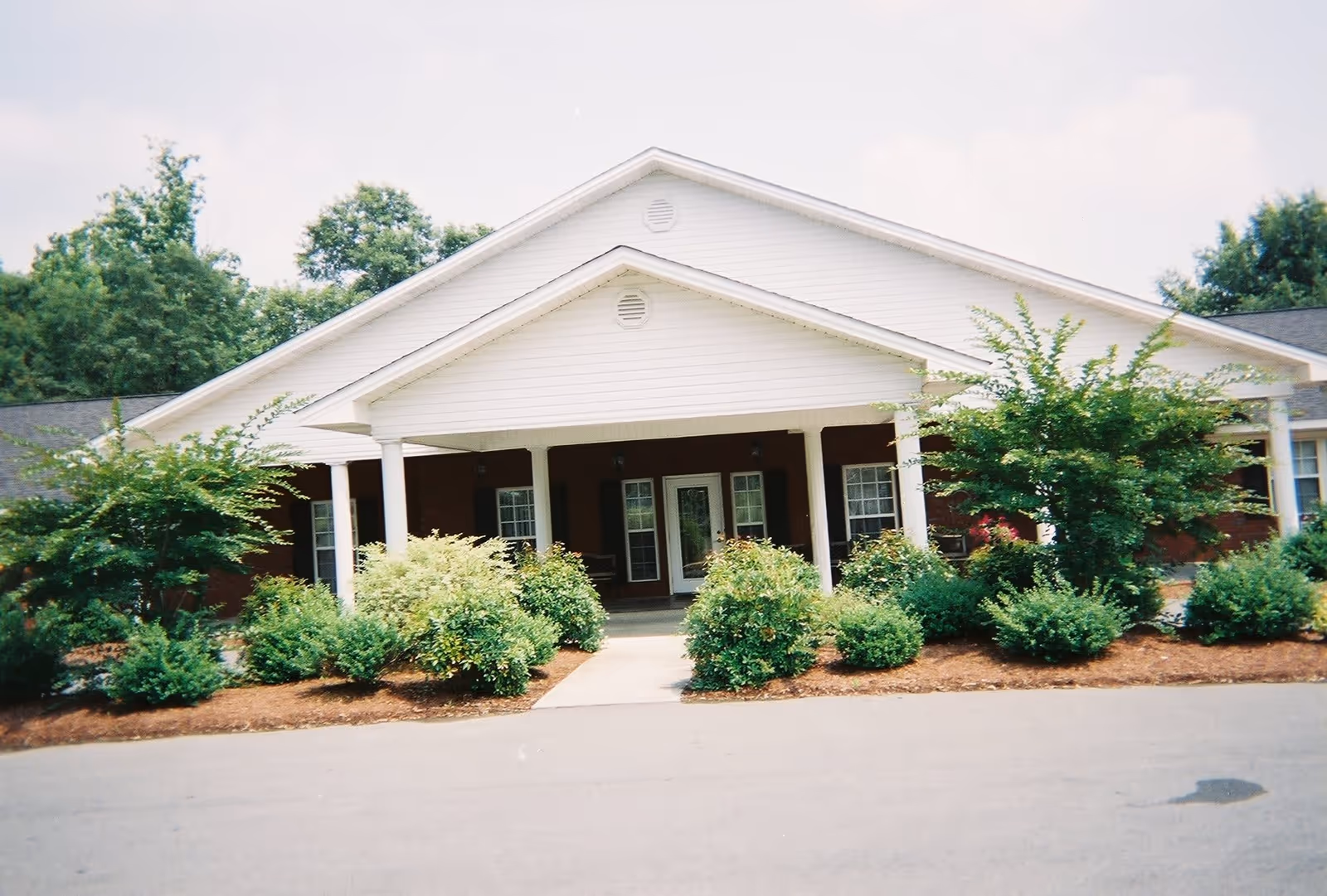 Front exterior view of a single-story assisted living facility building with a white gabled roof, white columns, and a covered entrance. The building is surrounded by neatly trimmed bushes and trees, with a paved driveway in front.