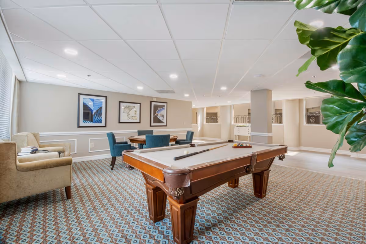 A well-lit recreational room featuring a pool table with a cue stick and billiard balls arranged on it. The room has patterned carpet flooring, beige walls with white trim, and a ceiling with recessed lighting. There are three framed pictures on the wall, a round table with four blue upholstered chairs, and a beige armchair with books on it. A large green plant is partially visible on the right side of the image.
