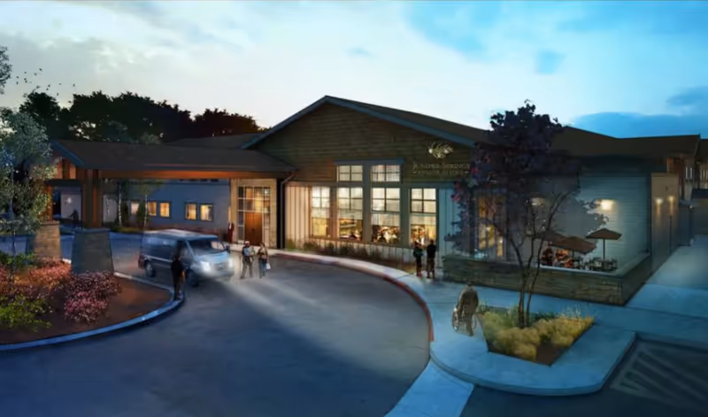 Front exterior of a senior living facility at dusk with a covered entrance, parked van, landscaping and people near the doorway.