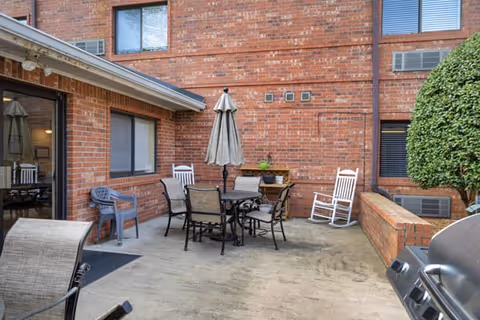 Outdoor patio area at Brookdale Fort Smith with a round table and six chairs, a closed umbrella in the center, two white rocking chairs, a gray plastic chair, a small wooden shelf with a potted plant, and a barbecue grill. The patio is surrounded by red brick walls and windows, with a neatly trimmed bush on the right side.