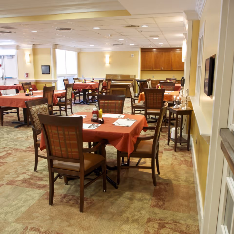 Dining room with multiple tables covered in red tablecloths, each set with napkins, utensils, ketchup, and mustard bottles. Chairs with patterned upholstery surround the tables. The room has beige walls, carpeted floor, and a piano against the far wall.