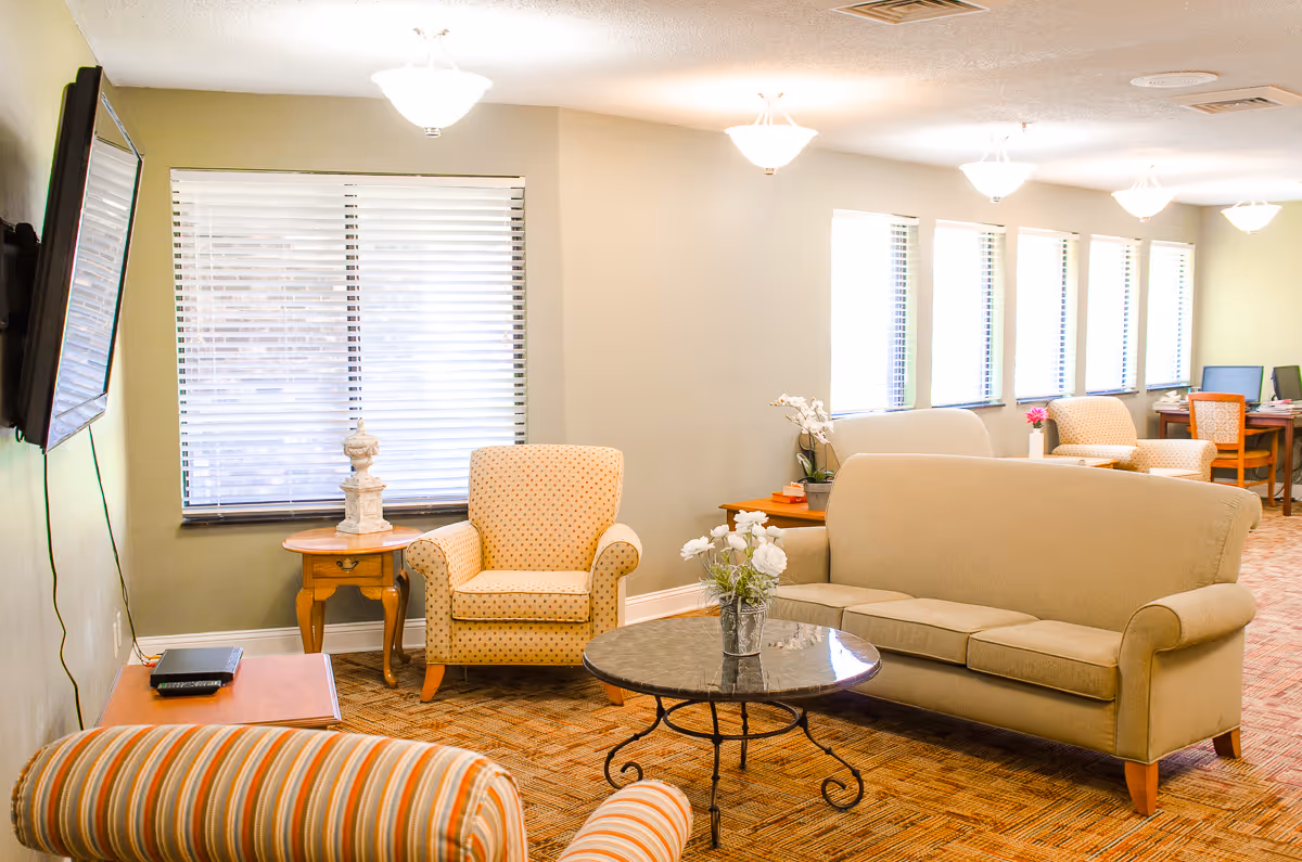 A bright communal living room with sofas, armchairs, a round coffee table topped with flowers, side tables and large windows.