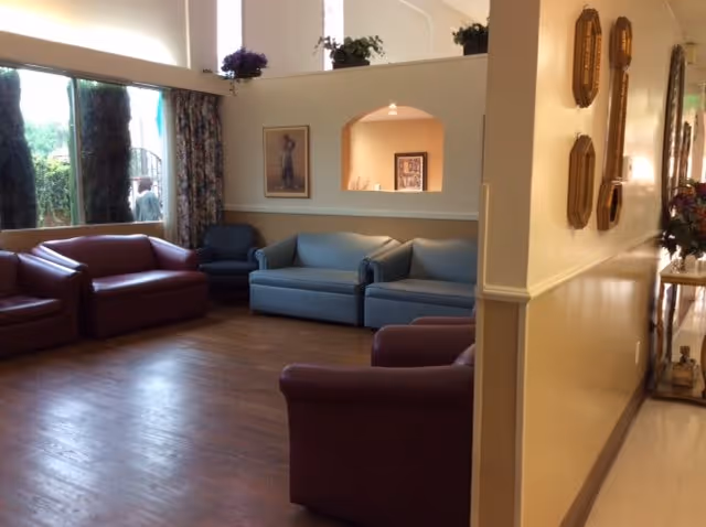 A living room area with multiple upholstered chairs and sofas in red and blue colors arranged around the room. There is a large window with floral curtains on the left side, and some framed artwork and plants on the walls and ledges. The floor is wooden, and the walls are painted light yellow and beige.