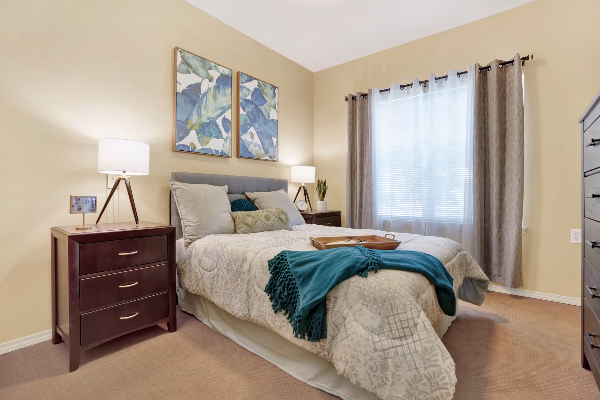 Well-lit furnished bedroom with a bed, two nightstands and lamps, framed artwork above the headboard, and a window with curtains.