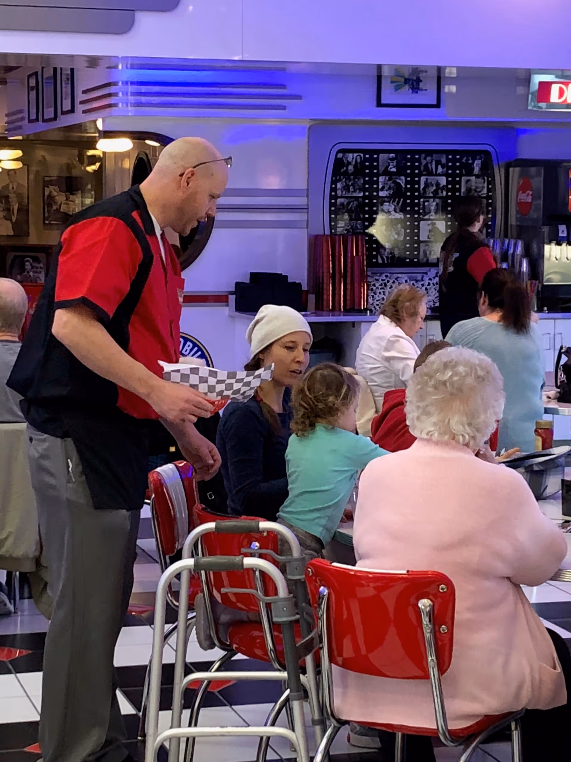 Interior diner-style dining area with a server in a red uniform attending to seated residents and visitors at red chairs and tables.