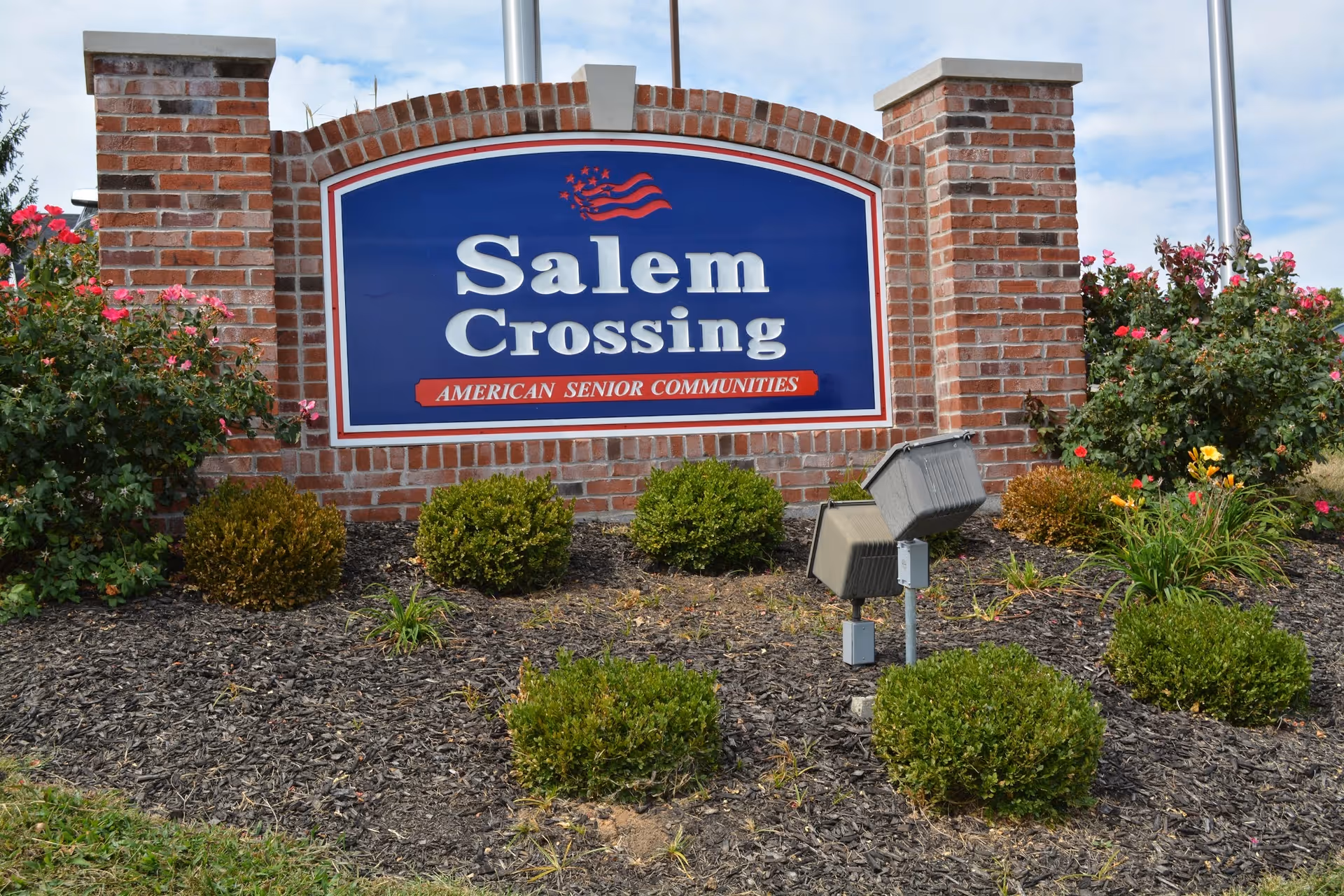 A brick sign for Salem Crossing, an American senior community, surrounded by landscaping with bushes and flowers under a partly cloudy sky.