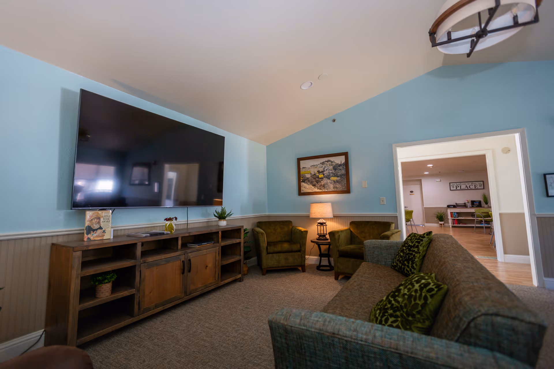 Cozy communal living room with a large wall-mounted TV, wooden media console, sofa and armchairs around a lamp.