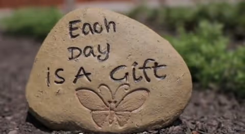 A decorative garden rock engraved with 'Each Day is a Gift' and a butterfly, sitting on mulch with greenery in the background.