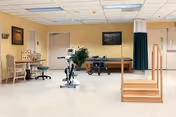 A spacious rehabilitation room with exercise equipment including a stationary bike and parallel bars with stairs for physical therapy. The room has beige walls, a light-colored floor, a wooden bench, a walker, and framed pictures on the walls.