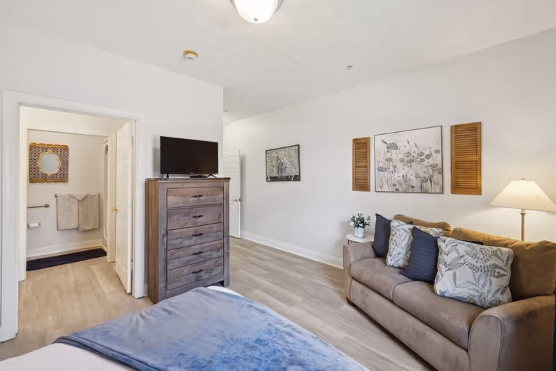 A cozy senior living bedroom with a bed covered in a blue blanket in the foreground, a wooden dresser with a TV on top, and a beige couch with decorative pillows and a floor lamp beside it. The room has light-colored walls and flooring, with framed artwork and wooden shutters on the wall. An open door reveals a bathroom with towels hanging and a decorative mirror.