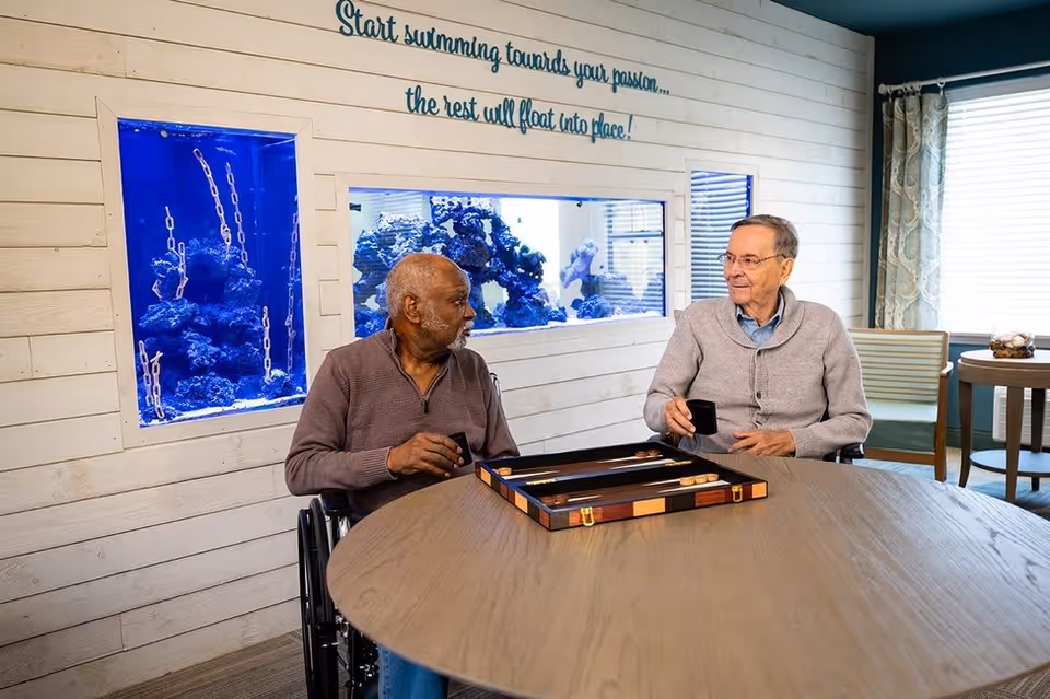 Two elderly men sitting at a round wooden table playing backgammon in a well-lit room with a large built-in aquarium in the wall behind them. One man is in a wheelchair, and both are holding small black cups. Above the aquarium, a sign reads, 'Start swimming towards your passion... the rest will float into place!'. The room has light-colored walls, a window with patterned curtains, and additional seating in the background.