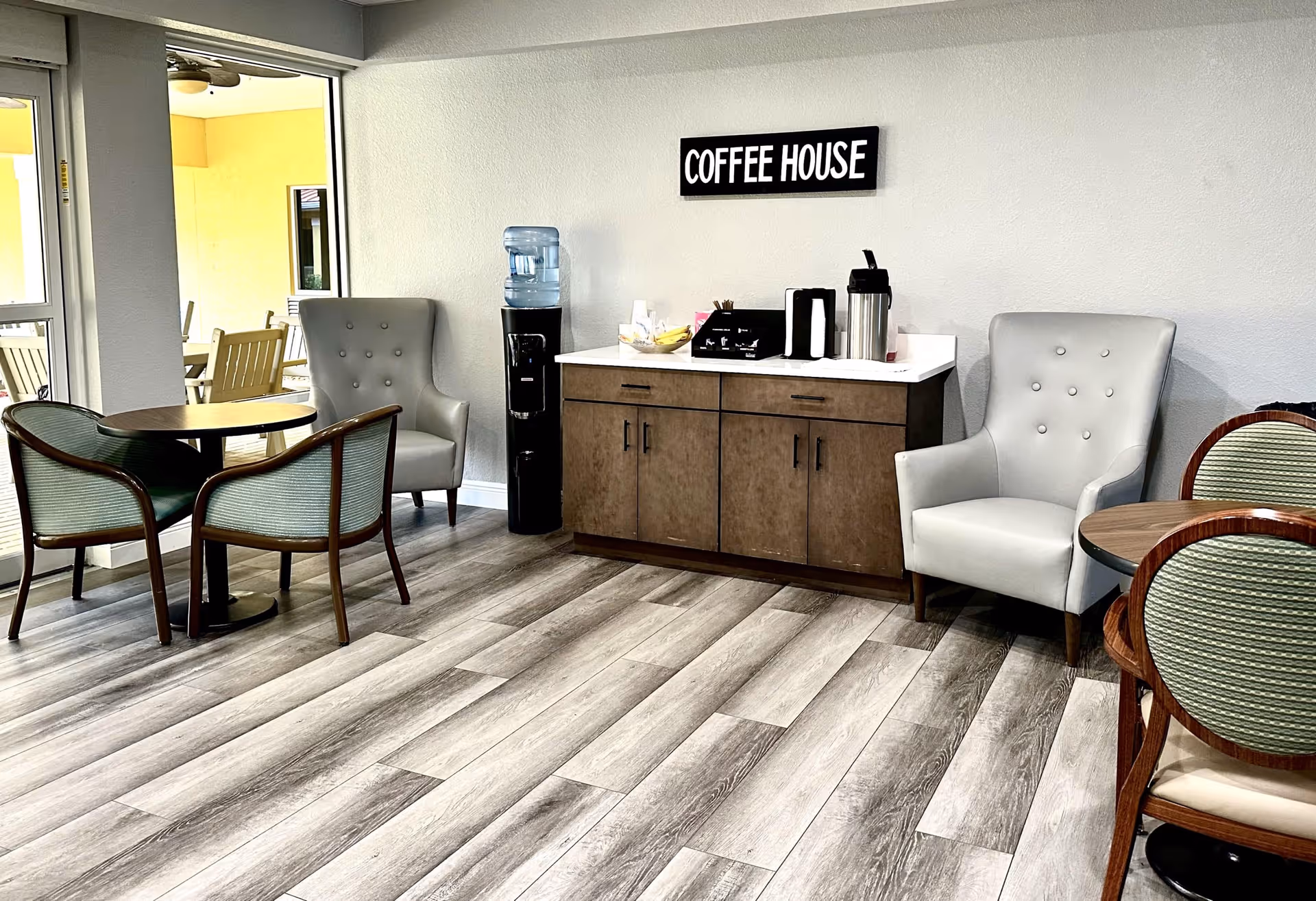 A cozy seating area in a senior living facility with a coffee station against a wall labeled 'COFFEE HOUSE'. The area includes two gray armchairs, two round tables with green cushioned chairs, a water dispenser, and a cabinet holding coffee supplies. Large windows allow natural light to enter the room.