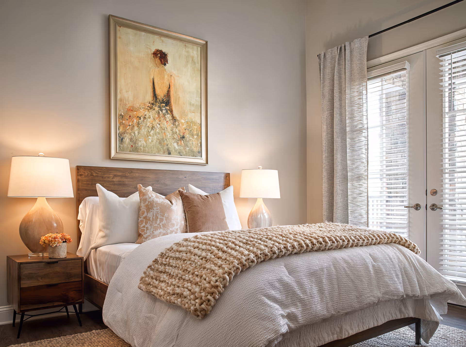 A cozy bedroom with a wooden bed frame, white bedding, and several decorative pillows. A chunky knit blanket is draped across the foot of the bed. Two bedside tables each hold a lamp with a beige shade, and a small flower arrangement is on the left table. Above the bed hangs a framed painting of a woman in a dress with her back turned. To the right, there are glass doors with blinds and light-colored curtains.