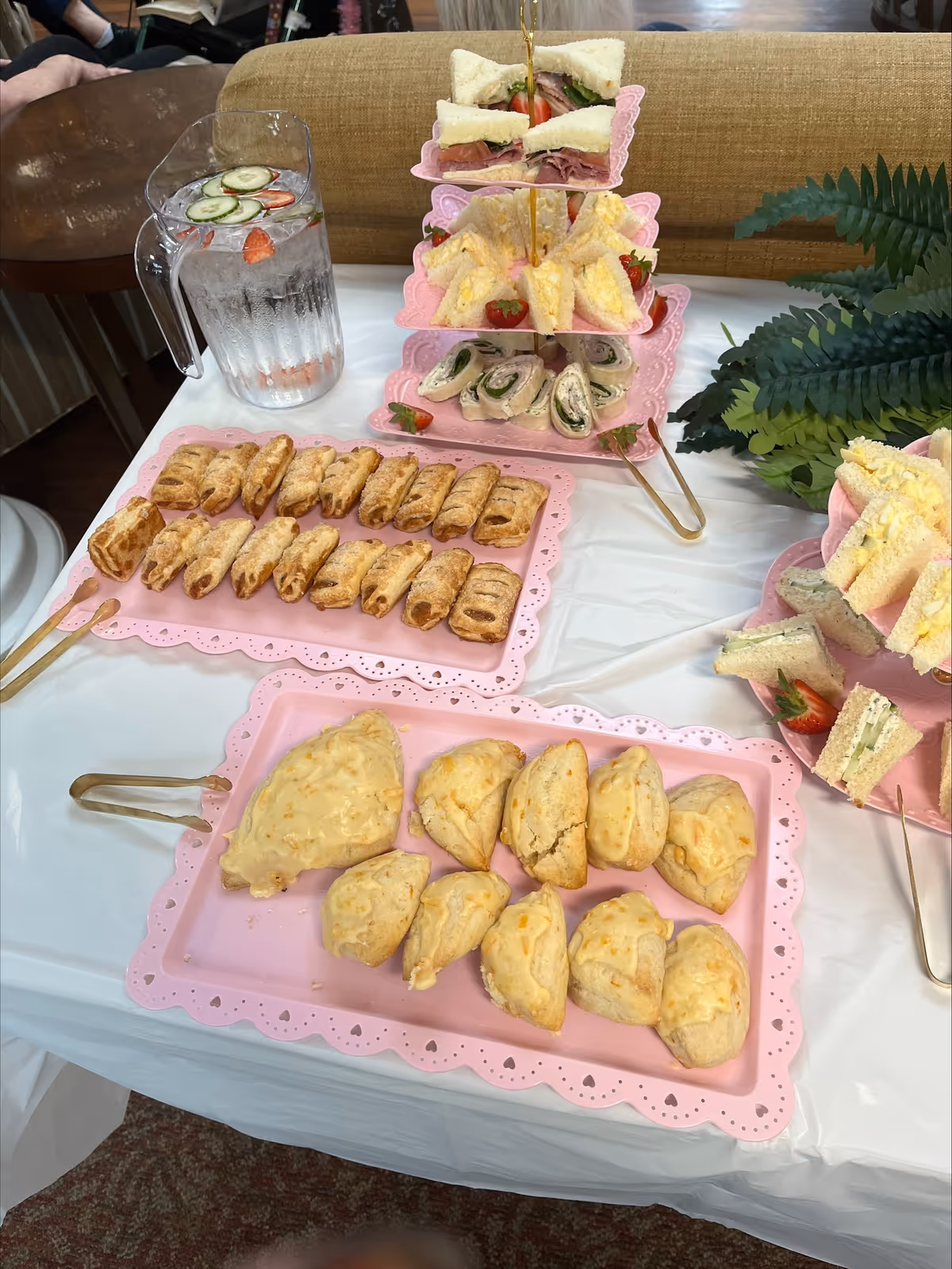 A table set with pink trays holding an assortment of finger foods including small sandwiches, wraps, pastries, and scones. There is also a pitcher of water with cucumber and strawberry slices. The table is covered with a white tablecloth and there is a green plant decoration on the side.