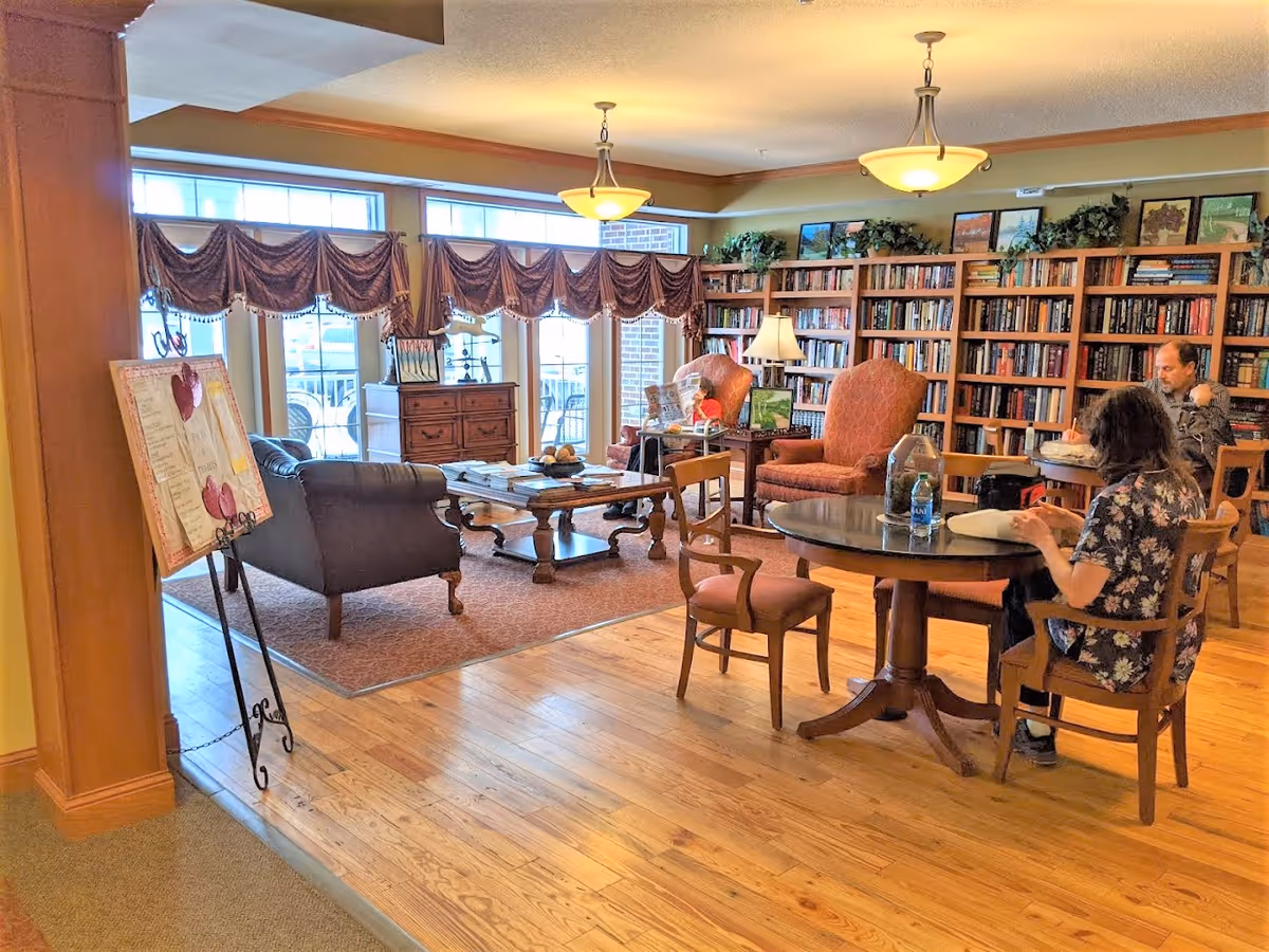 Comfortable library-style common room with bookshelves, armchairs, tables and large windows where residents are seated.
