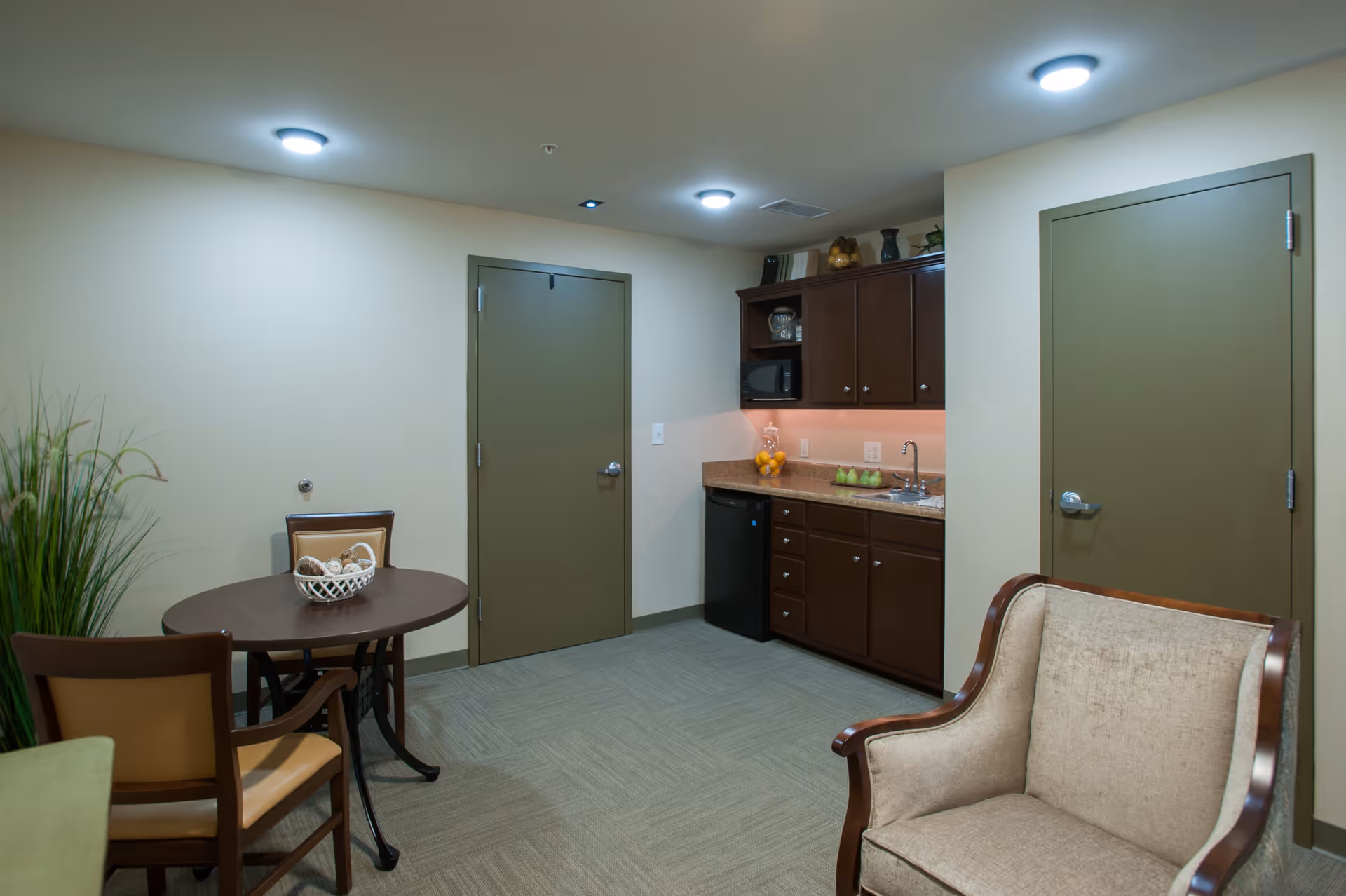 Interior room with a small kitchenette featuring dark wood cabinets, a countertop with a sink, a mini refrigerator, and a microwave. There is a round table with two chairs and a decorative basket on it, a beige upholstered armchair, and two closed olive green doors. The room has beige walls and carpeted flooring, with ceiling lights providing illumination.