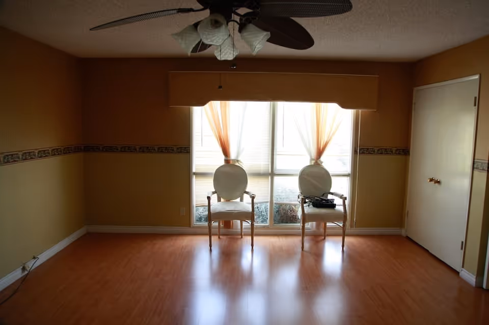 A mostly empty living room with two chairs facing a large window, wood laminate flooring, and a ceiling fan.