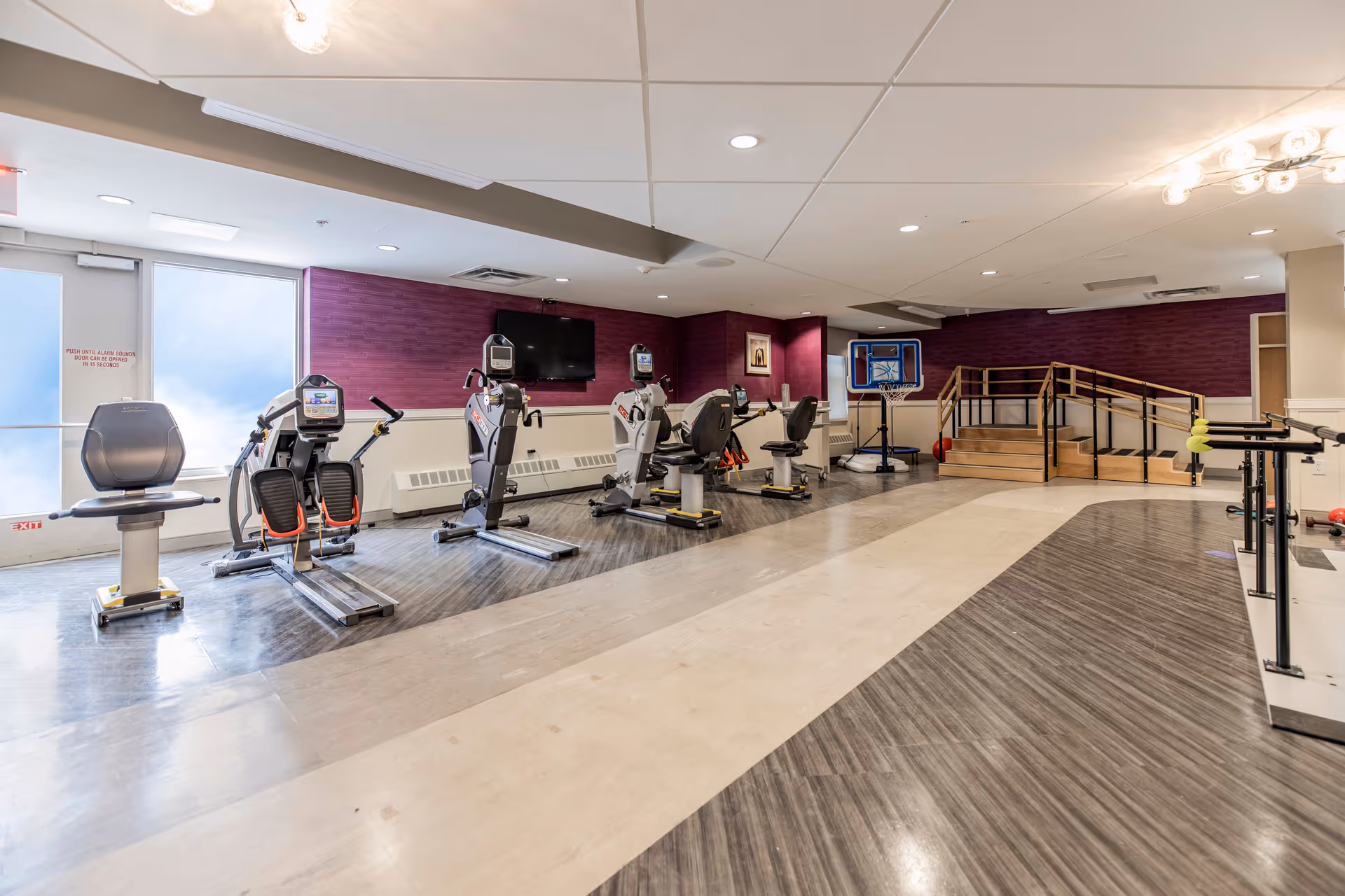 A rehabilitation and exercise room with several stationary exercise bikes and other fitness equipment. The room has a purple accent wall with a mounted flat-screen TV, a small basketball hoop, and a wooden ramp with handrails leading to a raised platform. The floor has a two-tone design with gray and beige colors, and the ceiling has recessed lighting.