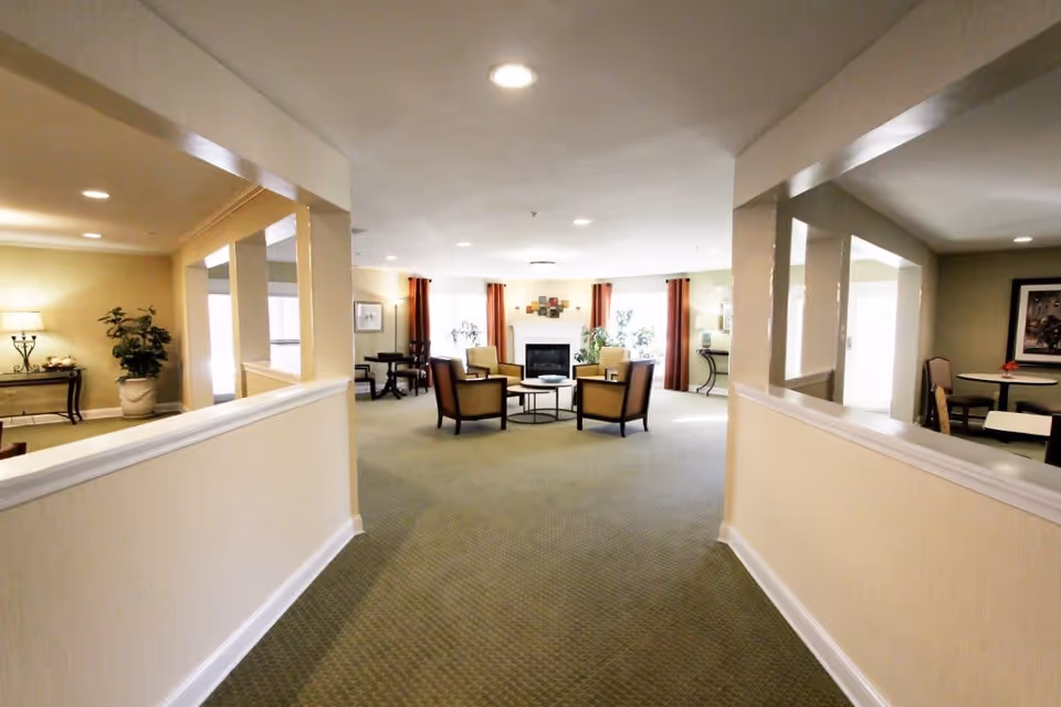 A spacious, well-lit common area in a senior living facility with a green carpet, beige walls, and recessed lighting. The room features a seating area with four chairs arranged around a small round table in front of a fireplace. There are large windows with red curtains, potted plants, and additional tables and chairs along the sides of the room.