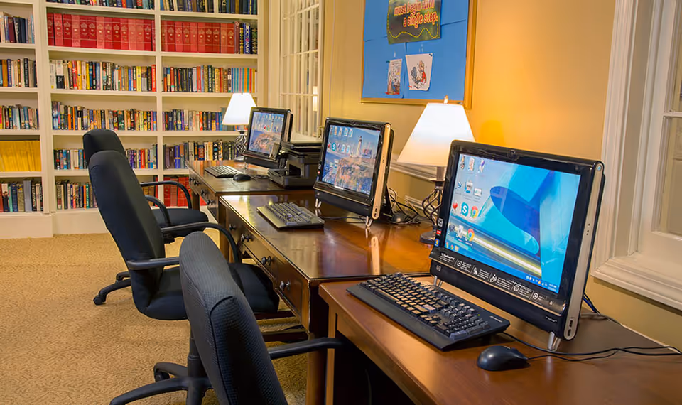 A computer room with three desktop computers on a long wooden desk, each with a keyboard and mouse. There are three black office chairs in front of the computers. Behind the desk is a bookshelf filled with books. Two table lamps provide warm lighting, and a bulletin board is visible on the wall.