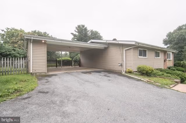 Exterior view of a single-story building with beige siding and a carport attached to the side. There is a paved driveway leading up to the carport, some green bushes near the building, and a wooden fence in the background with trees beyond it.
