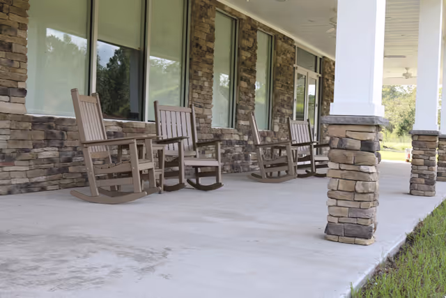 A covered outdoor patio area with four wooden rocking chairs lined up against a stone wall with windows. The patio has stone pillars supporting the roof and overlooks a grassy area.