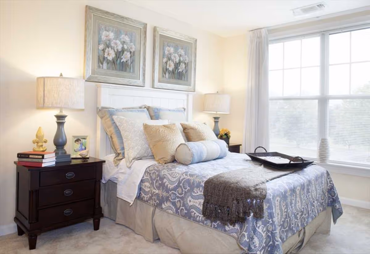 A neatly made bed with multiple pillows and a blue patterned bedspread in a bright bedroom. There are two framed floral paintings above the white headboard. On each side of the bed are dark wooden nightstands with lamps, books, and decorative items. A large window with white curtains lets in natural light.