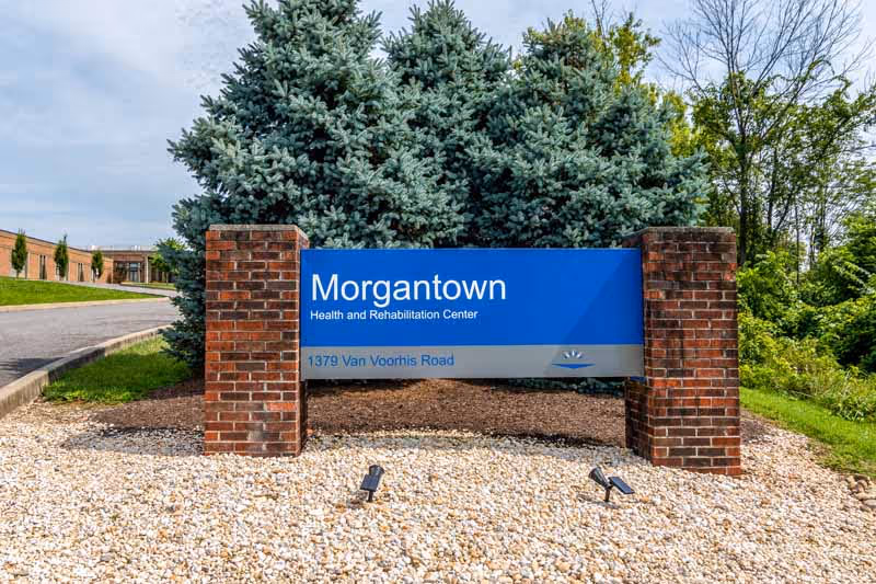Blue entrance sign reading 'Morgantown Health and Rehabilitation Center' mounted between brick pillars with trees and a driveway behind it.