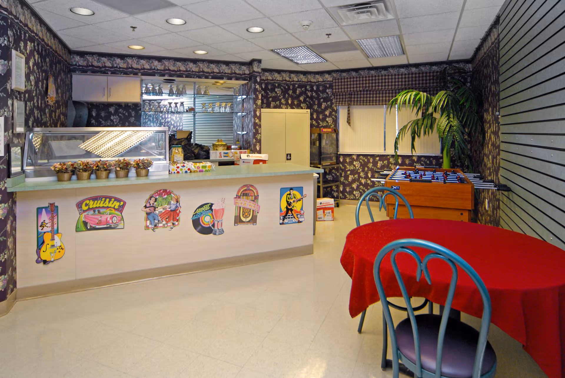A recreational room with a counter decorated with retro-themed stickers, small potted plants on the counter, a foosball table, a round table covered with a red tablecloth, and chairs with teal frames. The walls have dark floral wallpaper, and there is a large potted plant near the window with blinds.