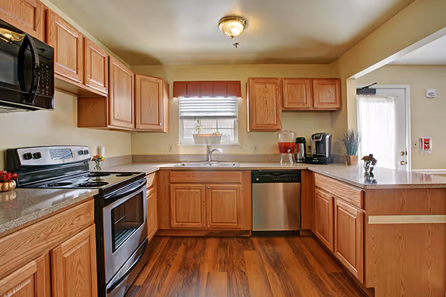 A spacious kitchen with wooden cabinets, a stainless steel oven and dishwasher, a microwave, and a coffee maker. The kitchen has a window above the sink with a small plant on the windowsill, wood flooring, and a light fixture on the ceiling. There is a door with a window and curtain on the right side.