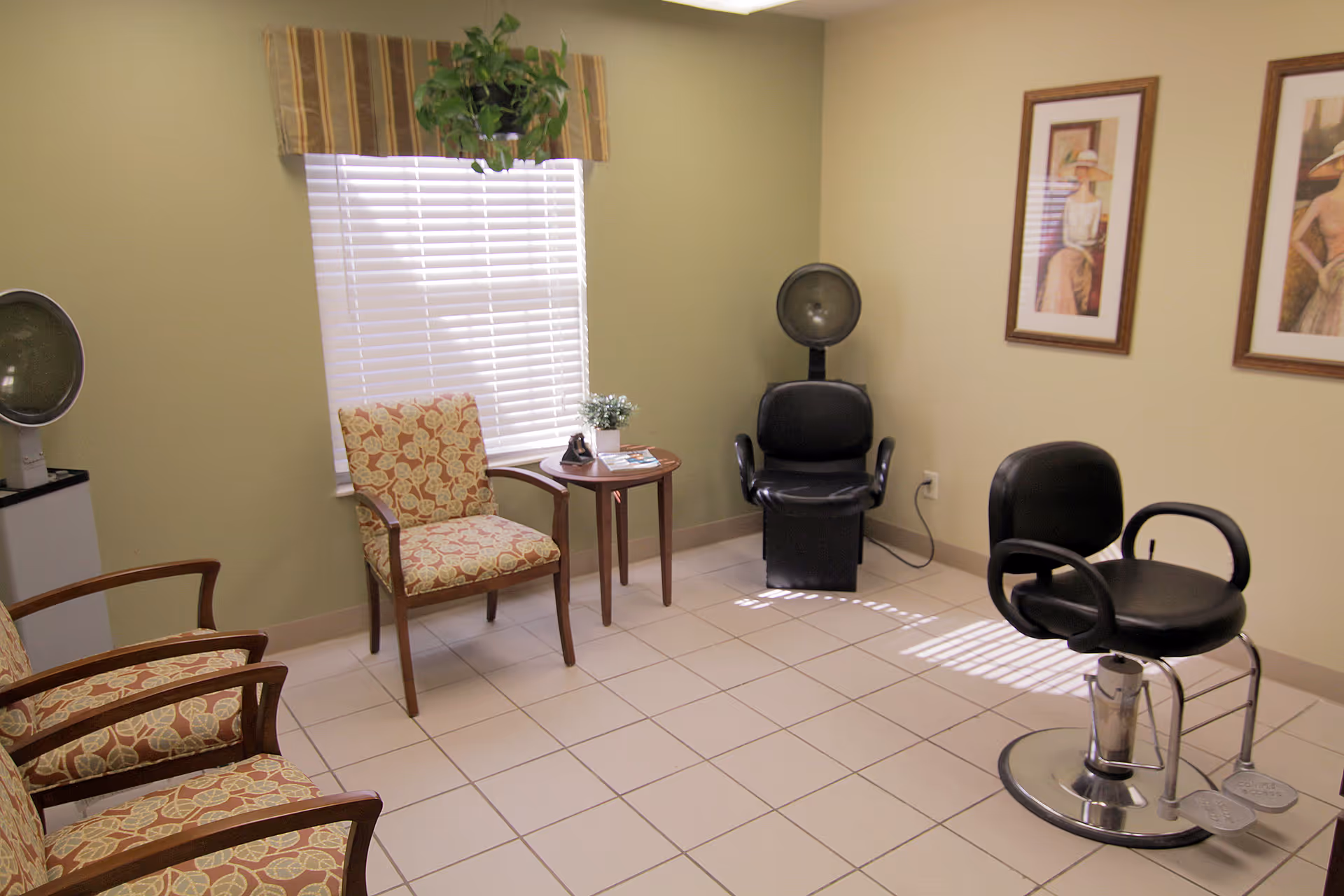 Small interior salon room with several chairs, a styling chair and hooded hair dryer, a window with blinds, and framed artwork on the walls.