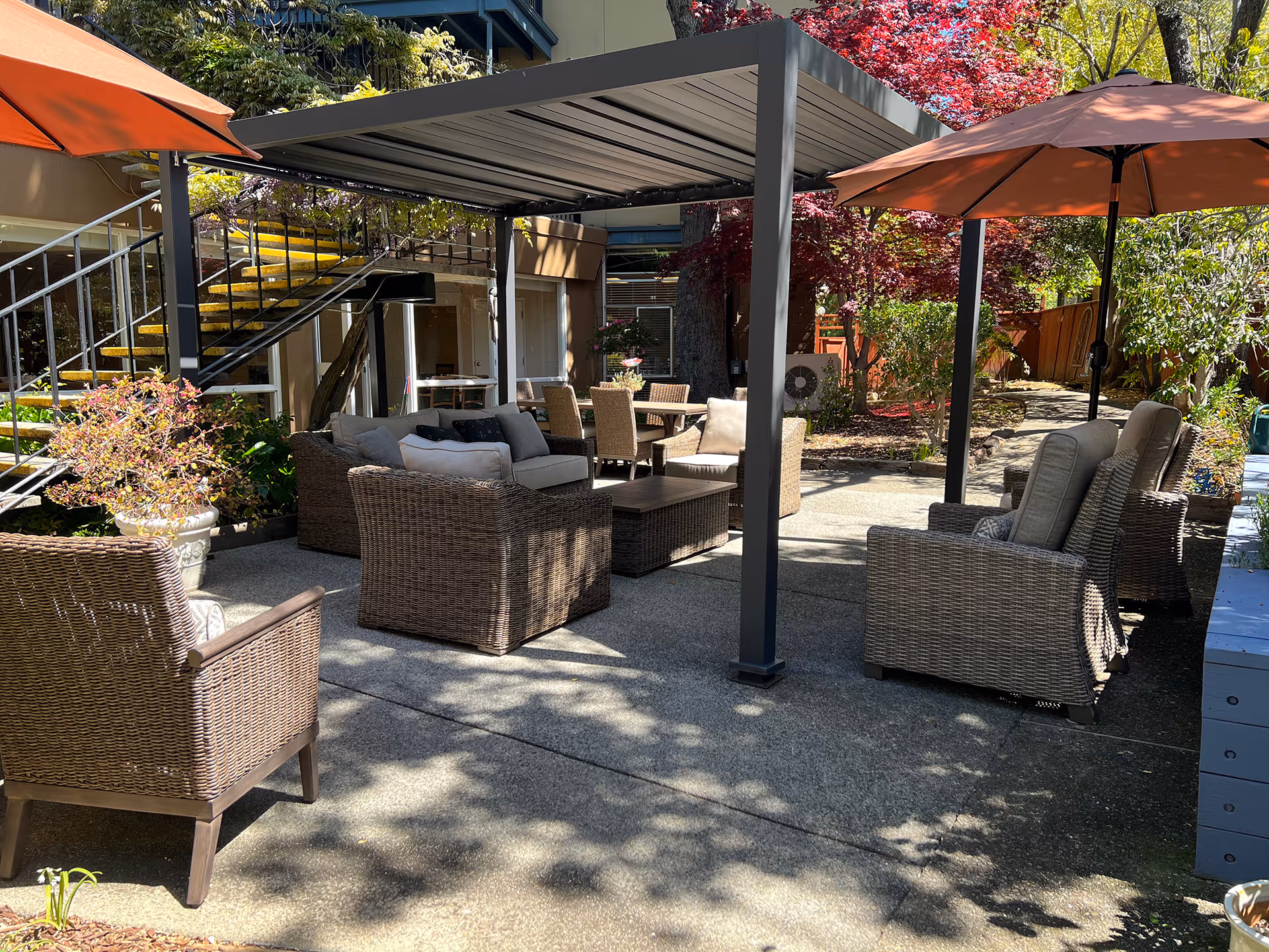 Outdoor courtyard with wicker seating, a pergola, umbrellas, and dining tables surrounded by plants and stairs.
