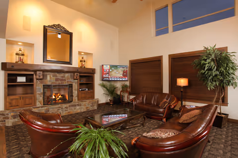 A cozy living room with a stone fireplace lit with a fire, surrounded by wooden shelves and a large mirror above it. The room features three brown leather sofas arranged around a glass coffee table, with a potted plant in the foreground and another tall plant near the window. The windows have wooden blinds, and a floor lamp provides additional lighting.