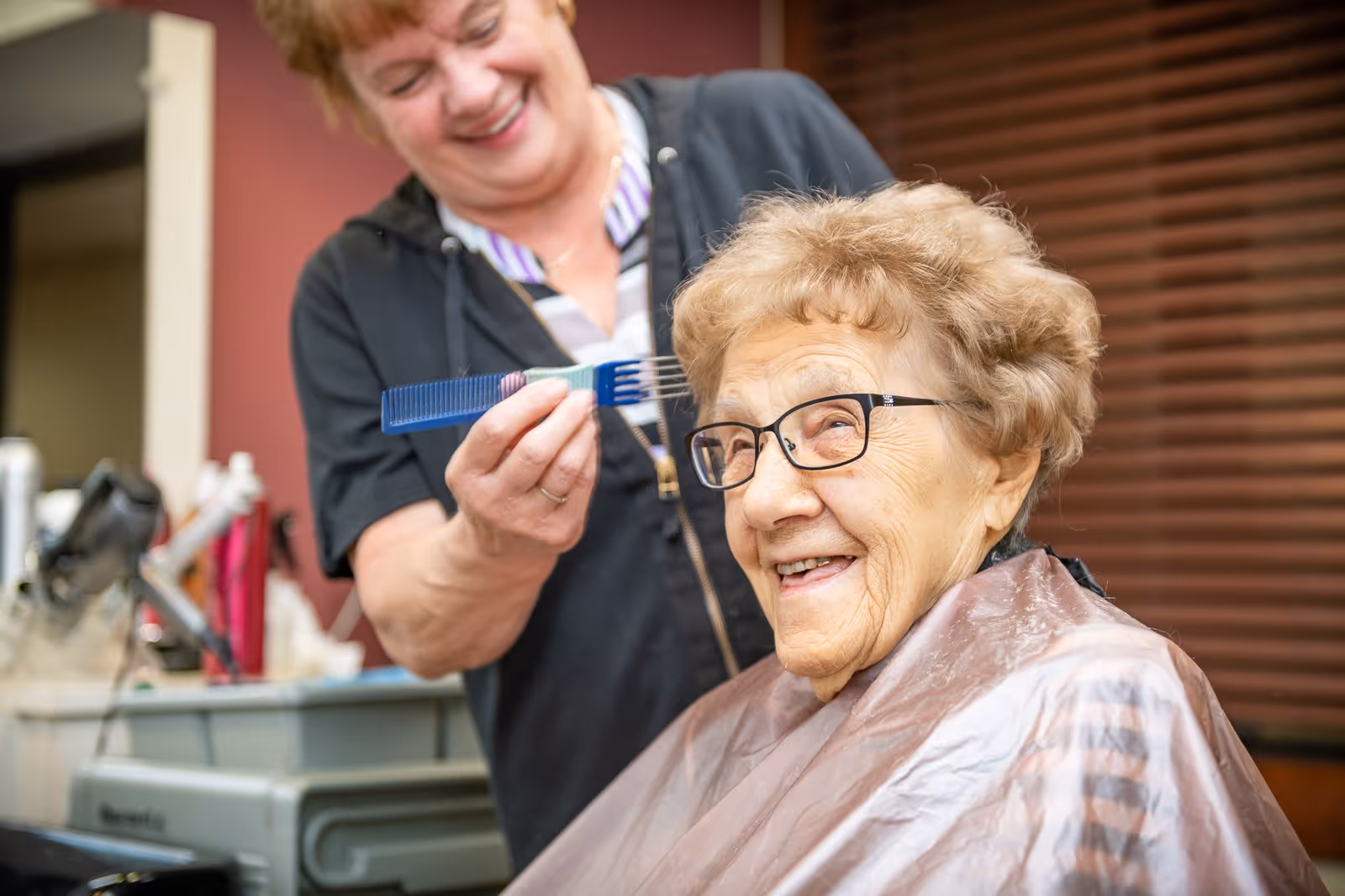 An elderly woman with glasses smiling while getting her hair styled by a hairdresser holding a comb in a salon setting.