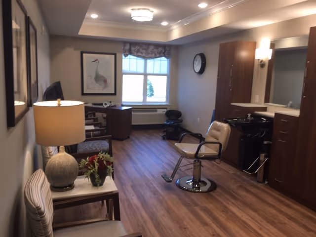 Interior salon-style room in a senior living facility with a styling chair and wash station, seating, a desk by the window, and wood flooring.