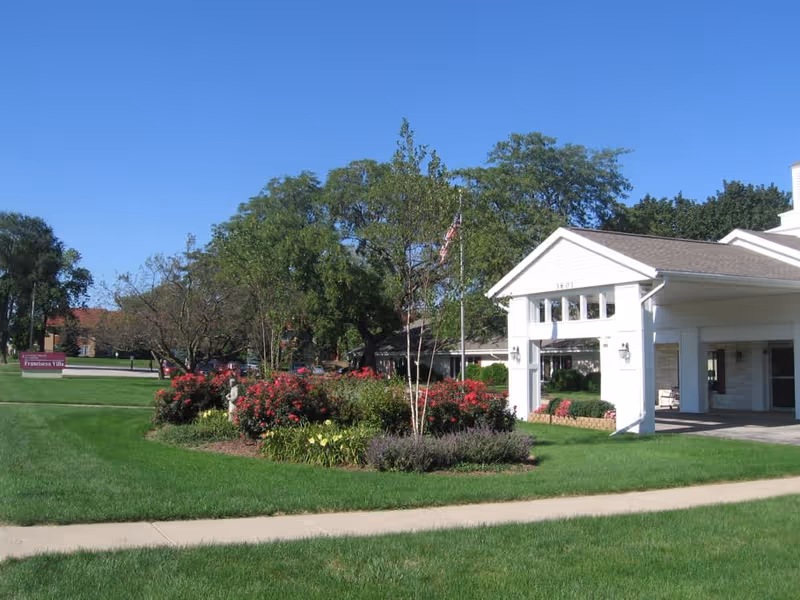 Exterior view of Franciscan Villa senior living facility showing a white entrance structure with a covered driveway, well-maintained green lawn, flower beds with red and yellow flowers, trees, and a clear blue sky.