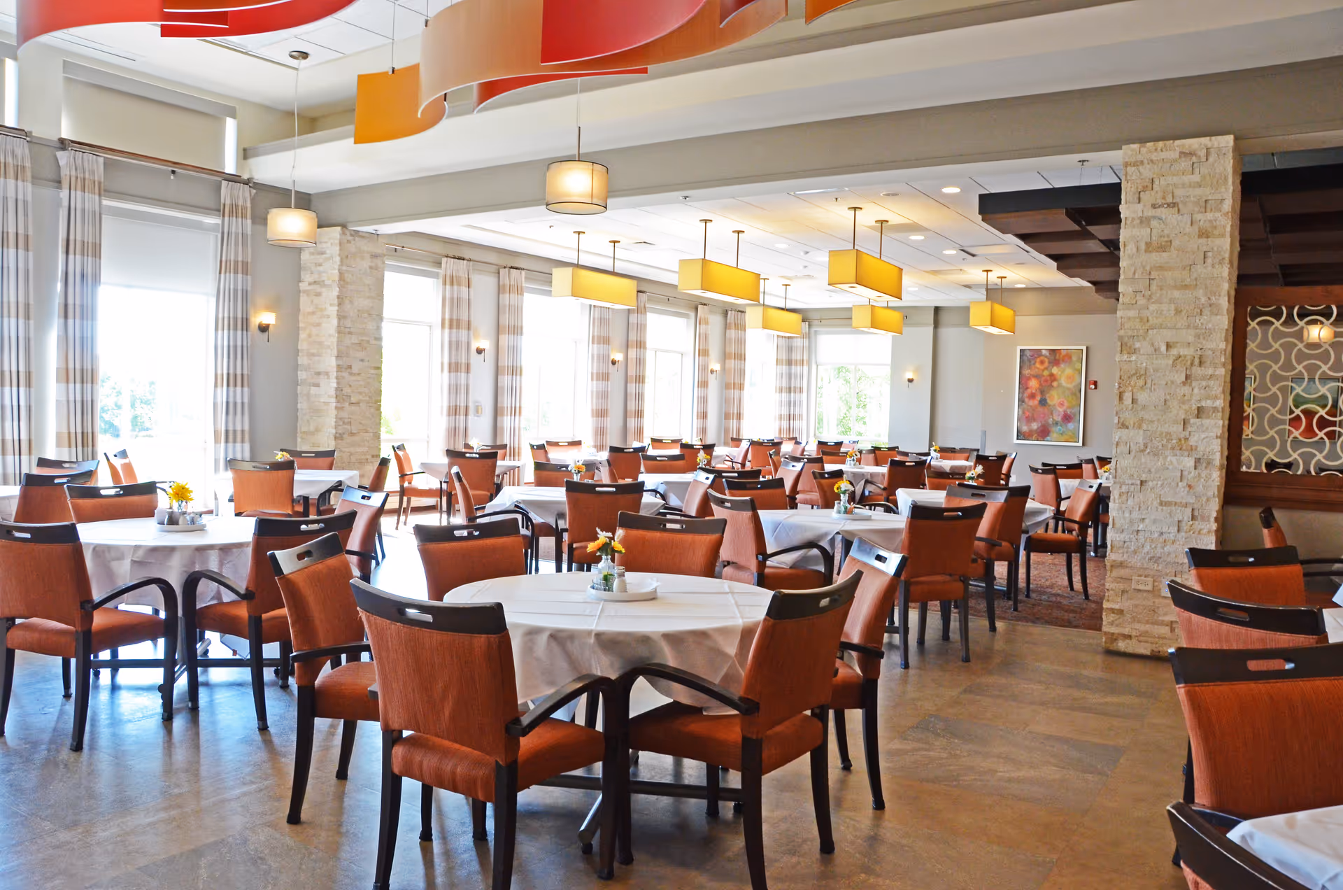 A bright and spacious dining room with multiple round tables covered with white tablecloths, each surrounded by orange cushioned chairs. The room features large windows with striped curtains allowing natural light to fill the space, modern pendant lights hanging from the ceiling, and decorative stone pillars. A colorful floral painting is visible on one wall.